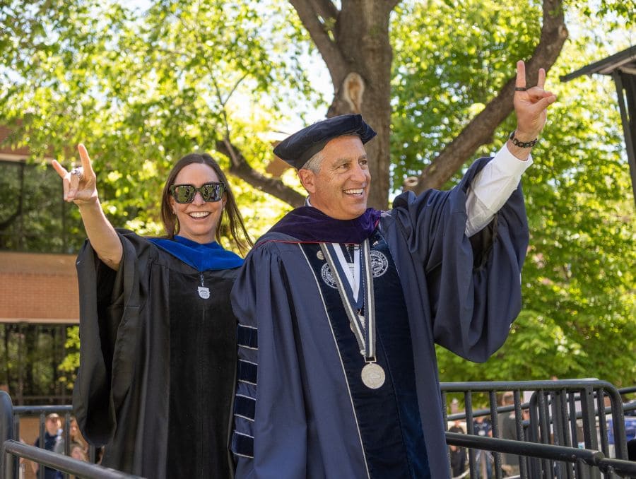 President Brian Sandoval and his wife, Lauralyn, wear academic regalia, smile and wave during an outdoor graduation ceremony.