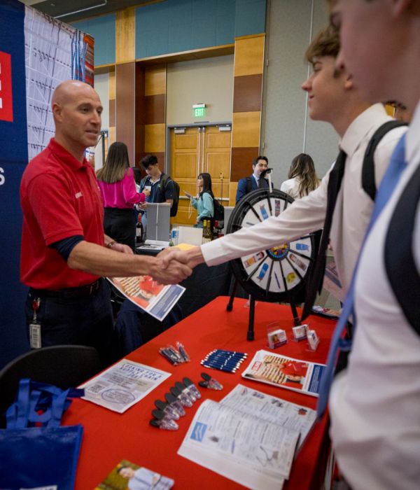 A man in a red shirt shakes hands with a young man in a white shirt at a career fair booth. A spinning wheel and promotional items are on the red table.