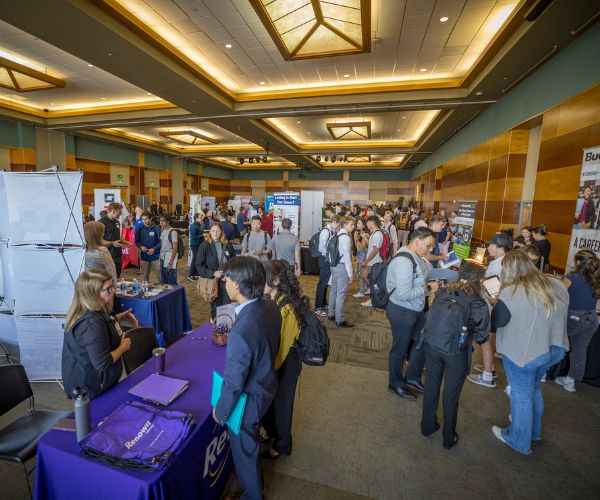 A bustling career fair in a spacious hall with attendees interacting at various booths. Tables are covered in colorful cloths, creating a lively atmosphere.