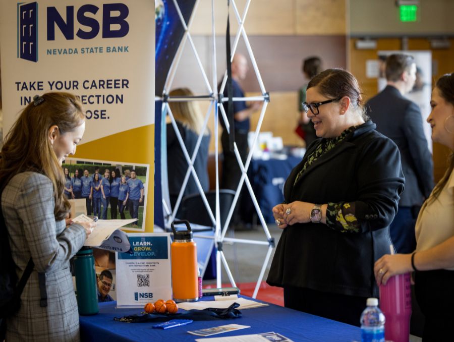 A career fair booth with a Nevada State Bank banner. Three women converse, one holding papers. A table displays promotional items and water bottles.