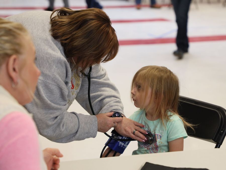 A healthcare worker in a gray sweatshirt checks a young girl's blood pressure in a clinic. The girl, in a light blue shirt, looks on attentively.