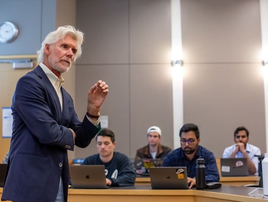 A man with gray hair lectures in a classroom, gesturing while four students with laptops listen attentively. 