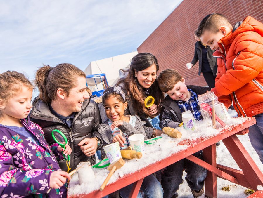 A group of kids and two adults are gathered around a snow-covered picnic table. They are wearing winter jackets, and engaging in a fun outdoor activity.