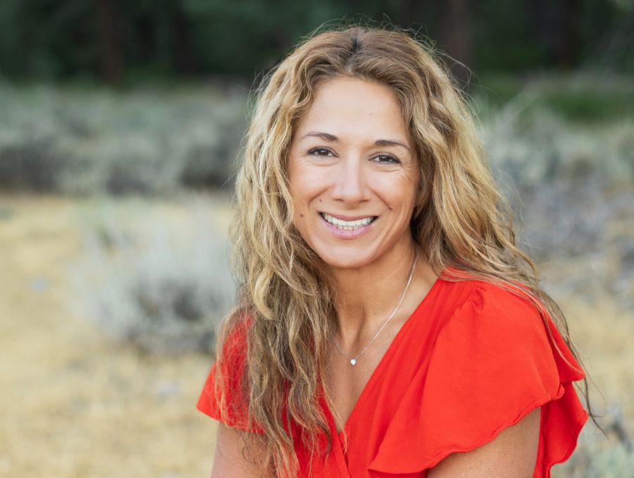 A smiling woman  wearing a bright red blouse. She is outdoors with blurred greenery and shrubs in the background.