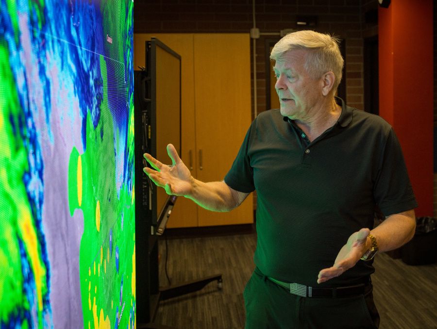 Man in a dark polo shirt gestures towards a large, vibrant digital map, displaying interest in its colorful forecasting data. 
