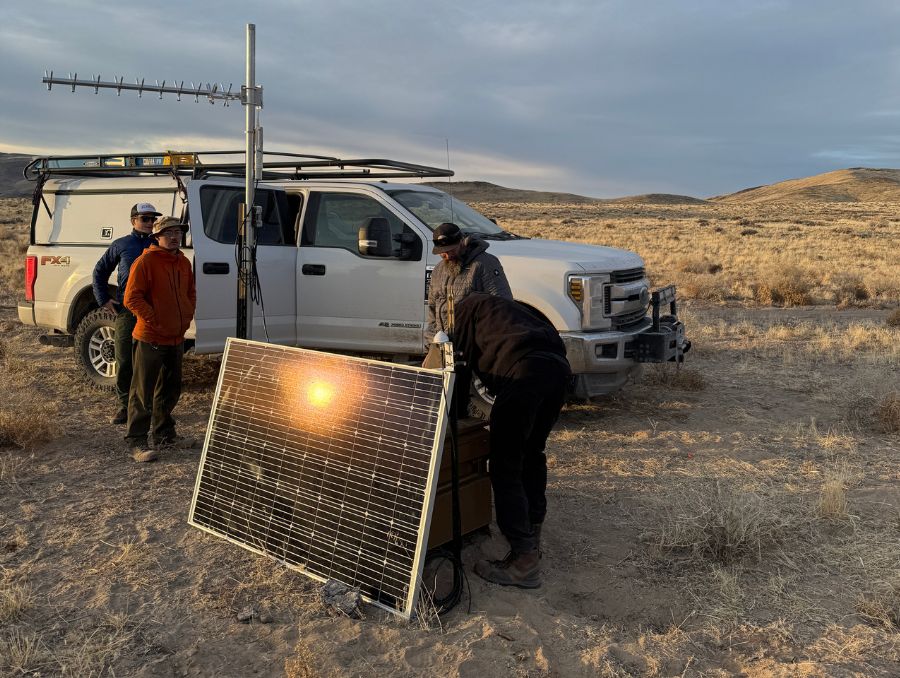 Three men stand in a desert landscape, positioned around a solar panel, showcasing renewable energy in a dry environment.