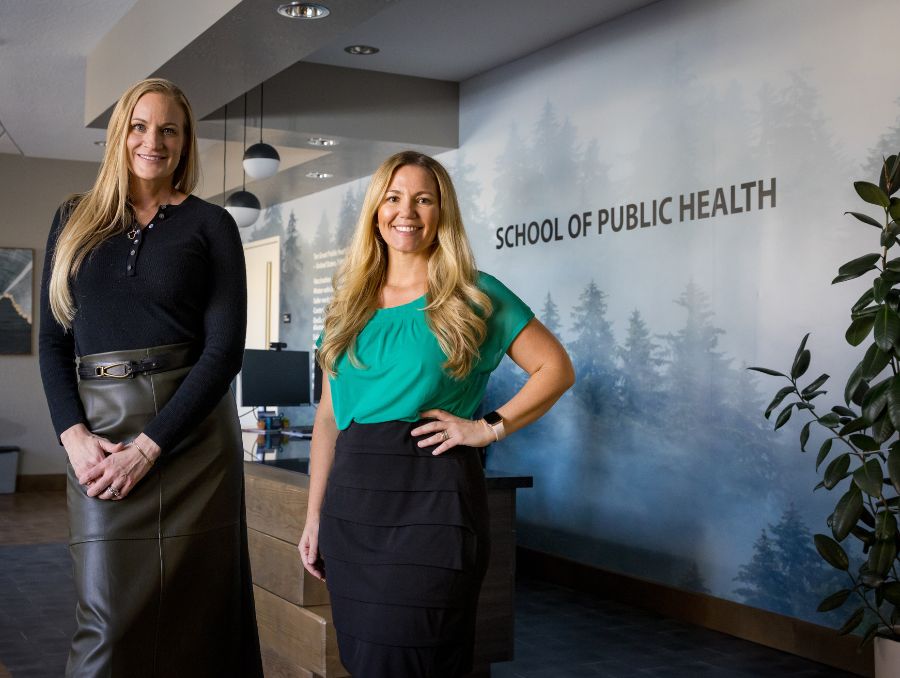 Two women pose in front of a sign reading "School of Public Health," smiling and dressed in professional attire.