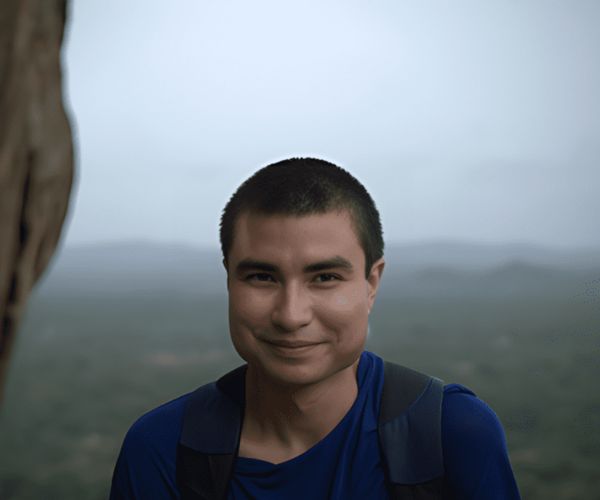 Jorge Esparza wearing a blue shirt and backpack smiles warmly with a blurred mountainous landscape in the background, conveying a sense of adventure.