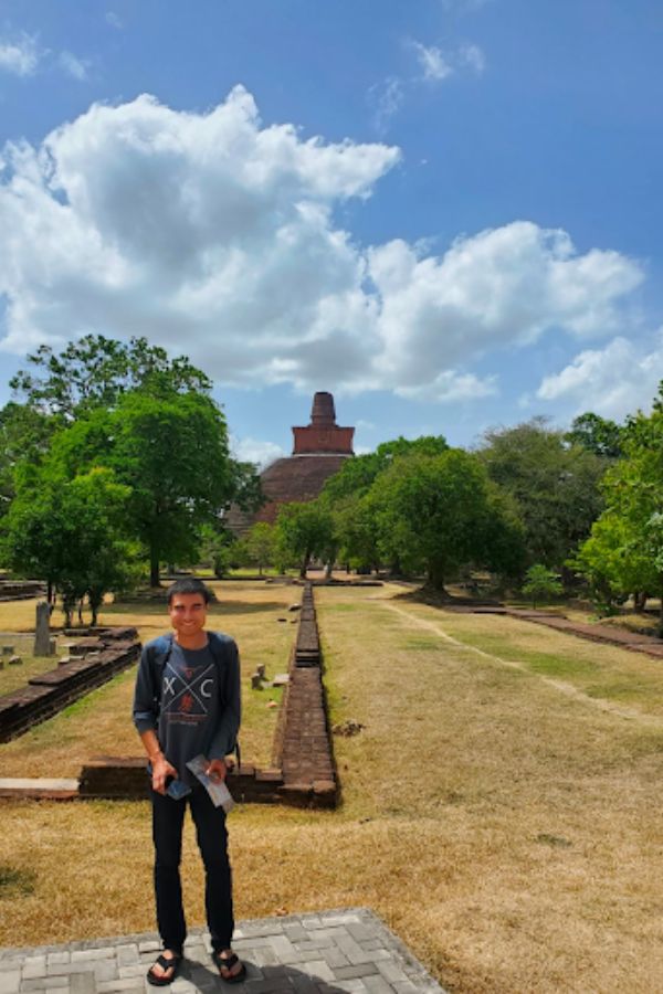 Jorge Esparza in casual clothes stands in front of an ancient brick stupa, surrounded by lush trees and a bright blue sky with fluffy clouds. Serene atmosphere.