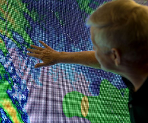 Man in a dark polo shirt gestures towards a large, vibrant digital map, displaying interest in its colorful forecasting data.