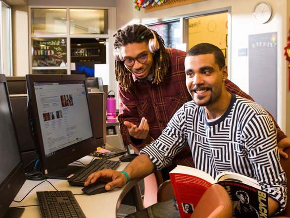 Two men sitting together with headphones on by a computer.