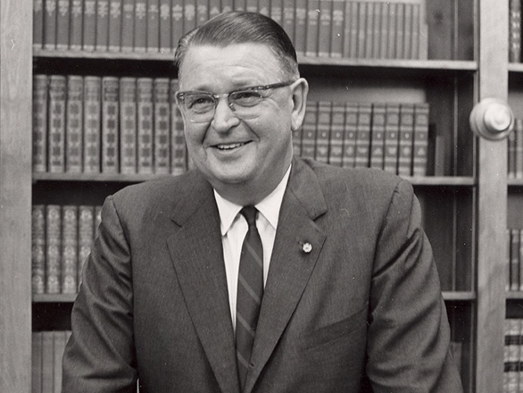 Black and white image of Charles Armstrong wearing a suit and resting his hands on a chair with a bookcase behind him