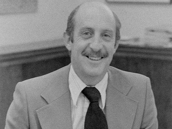Black and white image of Joe Crowley wearing a suit and standing in his office with a desk in the background