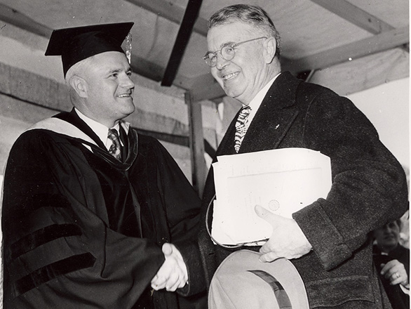 Black and white image of John Moseley dressed in academic attire shaking the hands of a gentleman carrying papers and a hat