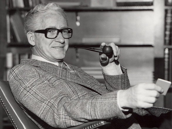 Black and white image of Max Milam seated in his office wearing glasses in a suit and holding a pipe to his mouth.