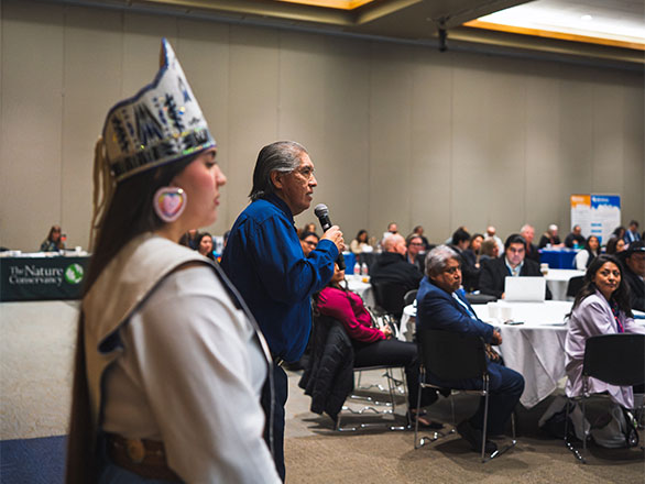 Tribal leaders speaking with a crowd seated at tables in a ballroom.