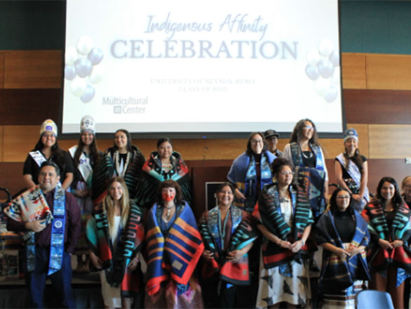 A group of students and community members pose together on a stage during an Indigenous Affinity Celebration at the University of Nevada, Reno. Many are wearing traditional clothing, patterned blankets, or cultural regalia, and some have ceremonial accessories. A large screen behind them displays the event title along with Multicultural Center branding.
