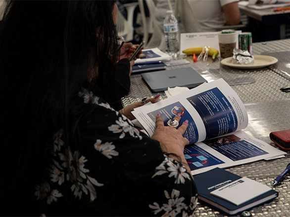 A person seated at a table flips through a printed booklet during a workshop or meeting. The table holds a laptop, notebook, pens, and refreshments, while another attendee in the background looks at a phone.