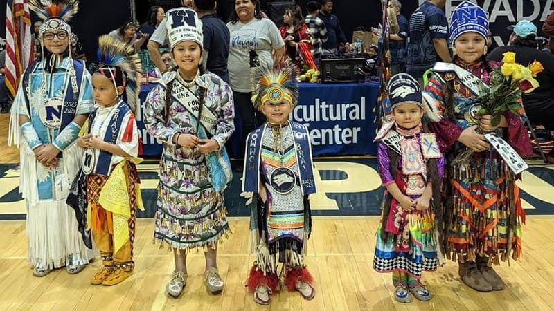 Five children wear traditional clothing for a powwow celebration, while other attendees stand behind them.