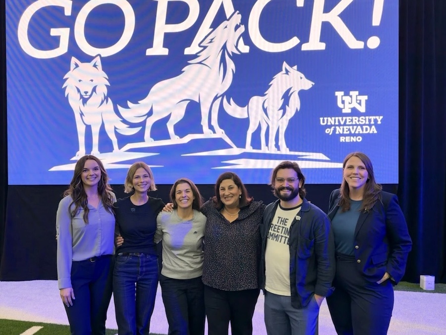 A group of people pose together in front of a screen that reads Go Pack! with the new regal trio of wolves.