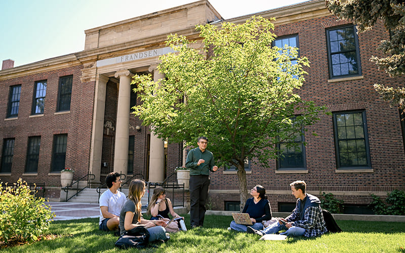 A professor conducts an outdoor class discussion with five university students seated on the lawn beneath a green tree outside the historic brick Frandsen building on campus.