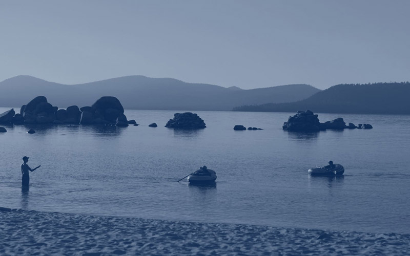 Calm waters of Lake Tahoe with people paddling near shoreline rock formations and mountain silhouettes, styled in a monochromatic blue duotone.