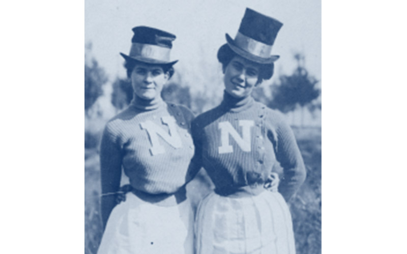 Archival black-and-white photograph treated with a navy duotone overlay, depicting two historic Nevada students standing side by side wearing top hats, long skirts, and sweaters with a block 'N' emblem.