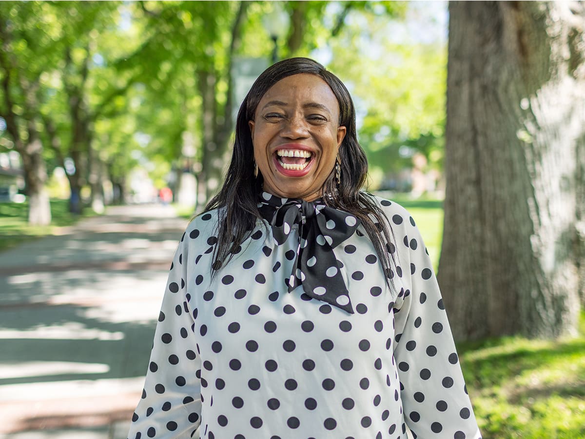 University faculty member in polka dot blouse laughing warmly while walking along the tree-lined Quad path