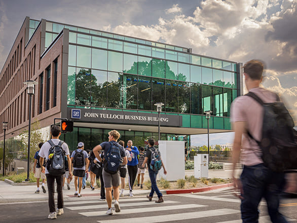 Students carrying backpacks cross the street toward the modern glass and brick John Tulloch Business Building on campus beneath a cloudy sky.