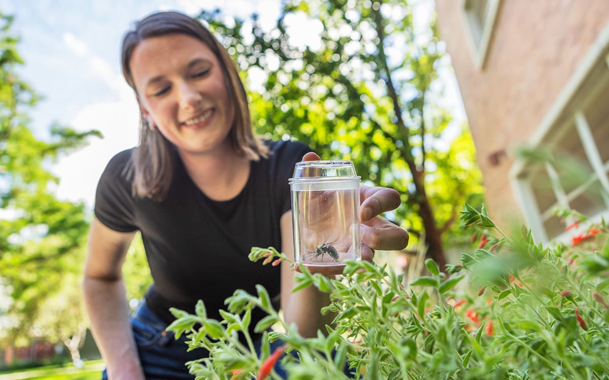 Student smiling while holding a clear specimen jar to examine a plant and insect sample outdoors