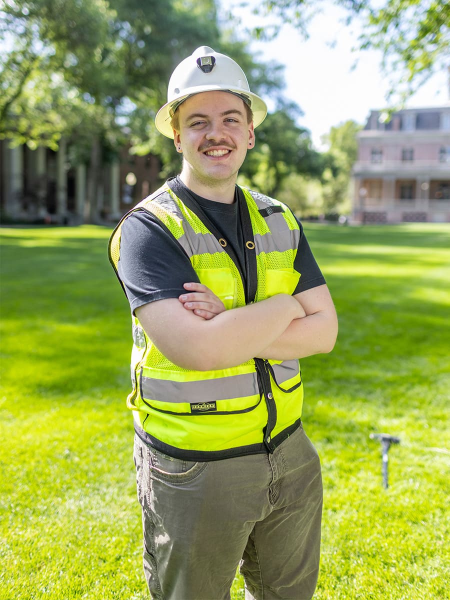 Engineering student wearing a safety hardhat and high-visibility yellow vest standing proudly outdoors on campus