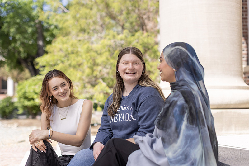 Candid outdoor portrait of three female university students conversing on a sunlit terrace next to a classical stone pillar, with one wearing a blue hijab and another in a navy blue University of Nevada crewneck sweatshirt against green campus trees.