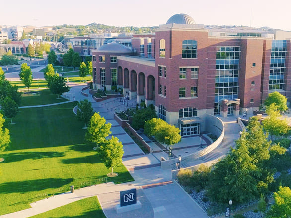 Aerial view of the University of Nevada, Reno campus featuring the Mathewson-IGT Knowledge Center in the foreground, a large red-brick building with arched windows and a domed roof. Surrounding the building are green lawns, tree-lined walkways, and landscaped paths leading toward other campus structures under a clear blue sky.