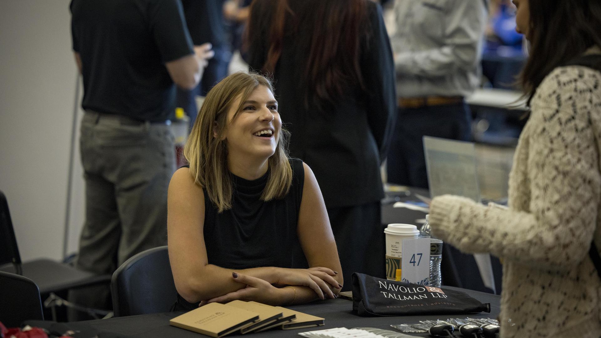 A woman sitting behind a table at a fair on campus with free promotional items scattered in front of her for the taking.