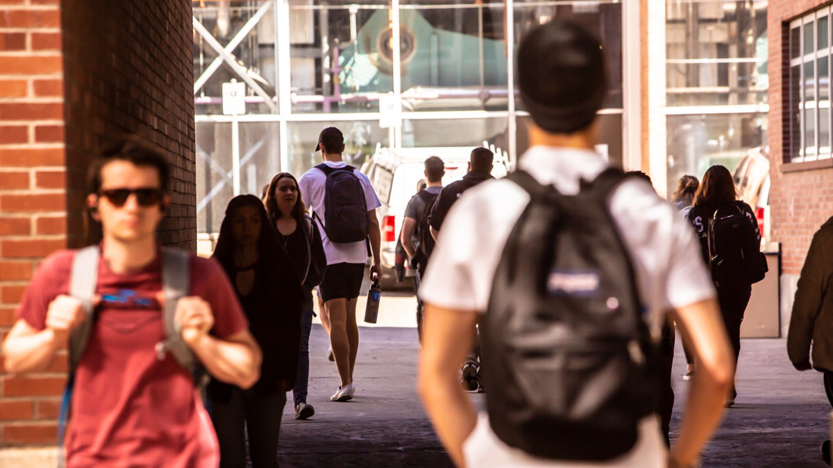 Students walk under a pedestrian walkway on campus, many wearing backpacks