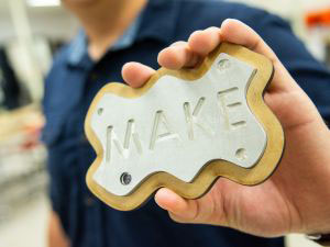 Close-up of a hand holding a metal sign mounted on wood with "MAKE" engraved in the metal