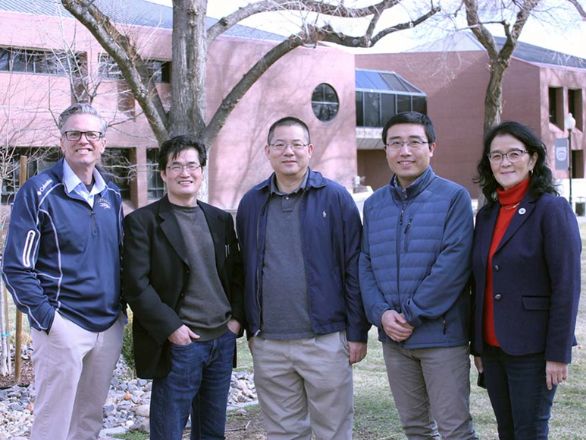 Four men and one woman stand in a line in front of a grassy area, trees and a red brick building.