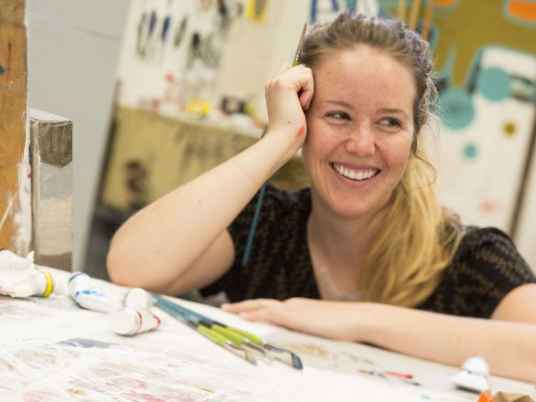 Student sitting at a table with art supplies on the table and paintings on easels in the background
