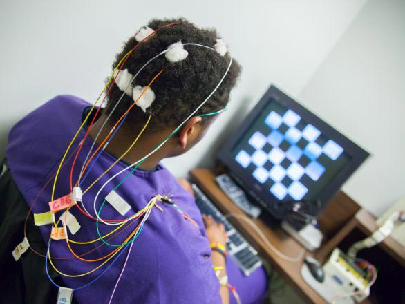 View of the back of a research participant with sensors attached with colorful wires to head and looking at a computer
