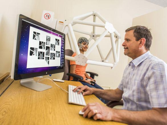A faculty researcher at a computer looking at a live video image with a participant sitting inside test equipment with sensors on their head