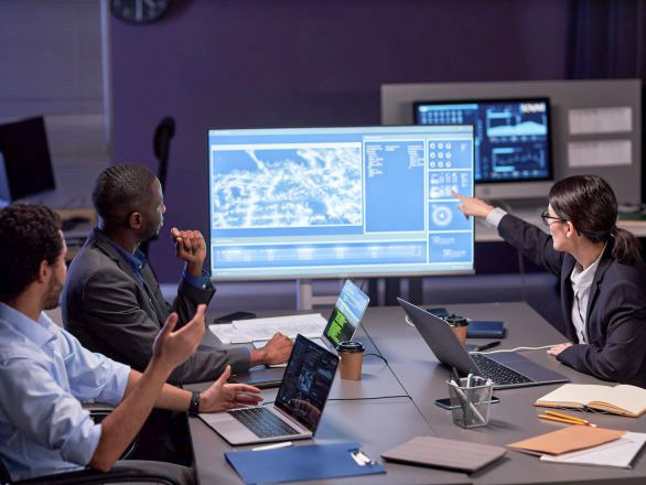 Three people dressed in business clothing working at a table with laptops and a large monitor displaying chart graphics.