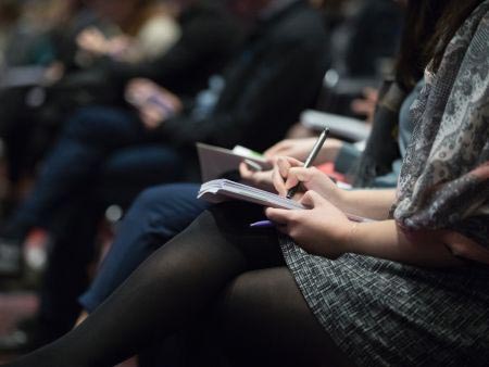 Close-up of a person writing notes sitting at a conference in a row of people (just showing person's lap)