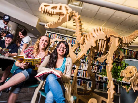Two students sitting in the education building library with a large, wooden dinosaur skeleton art piece in the background