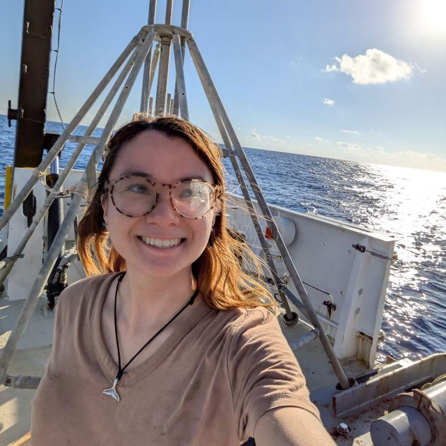 Hiroka stands smiling on the deck of the research vessel with the ocean and sun behind her.