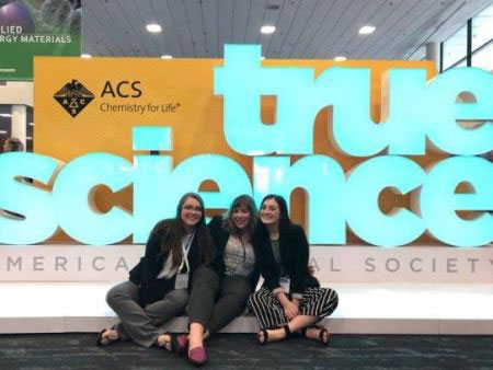 Three students sitting in front of a sign reading "true science" at the American Chemical Society conference