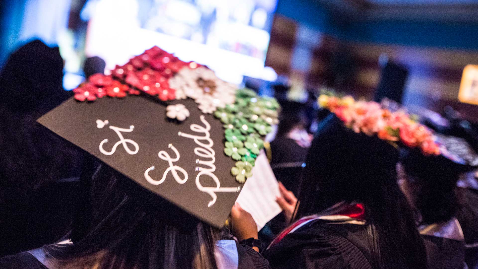 Students sit in graduation attire at commencement with one mortar board in focus with the words "Si Se Puede" written on it.