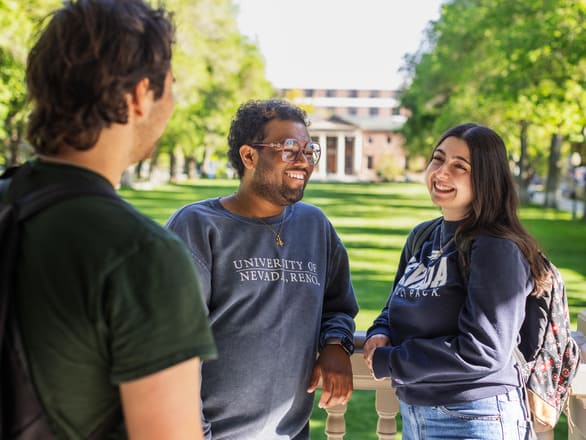 Three individuals stand outdoors on a sunny day, engaged in conversation near a railing with a green lawn and trees in the background. Two wear sweatshirts with visible text: one reads “University of Nevada, Reno,” and the other reads “Nevada Wolf Pack.” All have backpacks, suggesting a campus setting, and a large academic building with columns is visible in the distance.