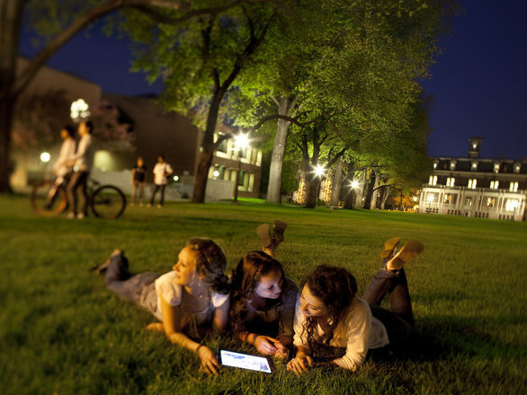 A group of three students lie on the quad at the University of Nevada, Reno in the evening and look at a laptop together.