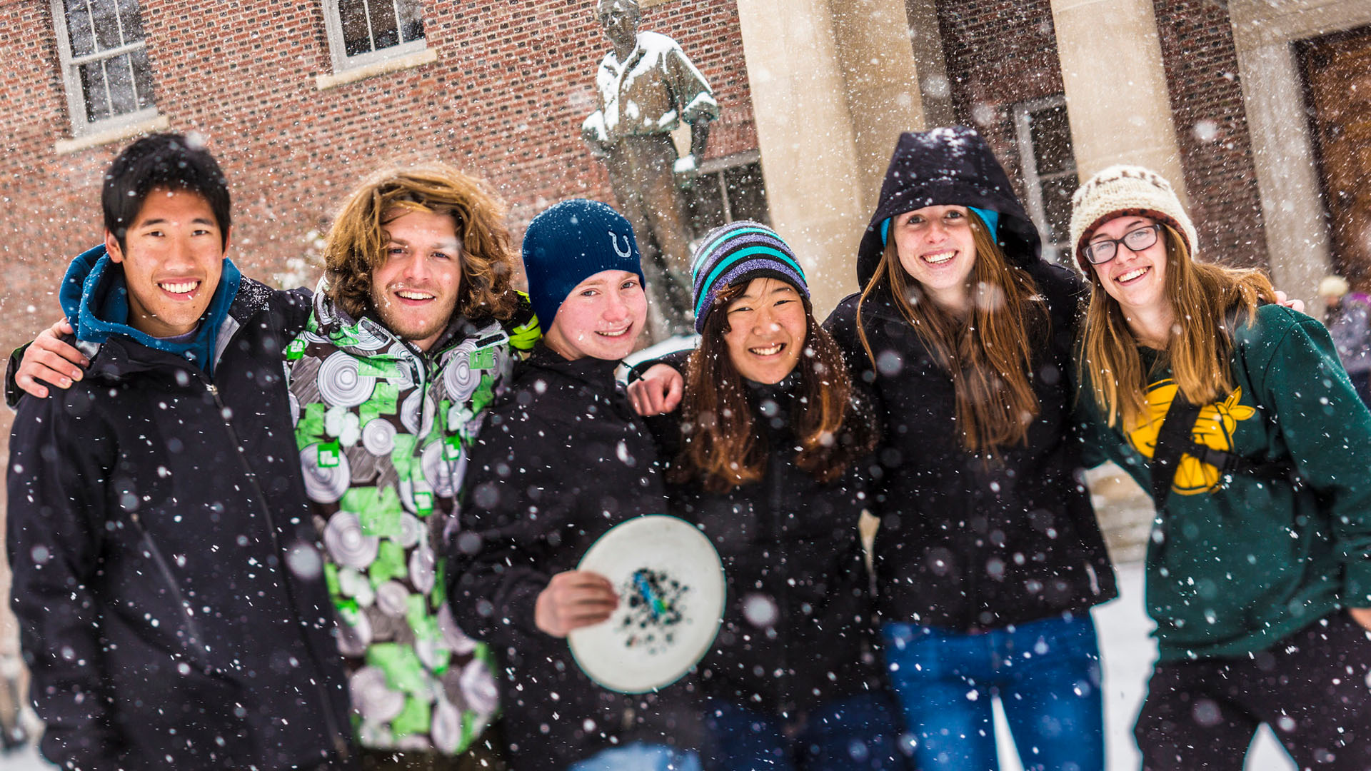 Students in winter gear gather on the University quad, iwth a frisbee, as puffy snow falls around them.