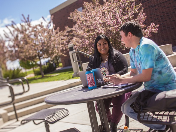 Two students sit at a table outside the Joe Crowley Student Union with trees blooming in the background on the campus of the University of Nevada, Reno.